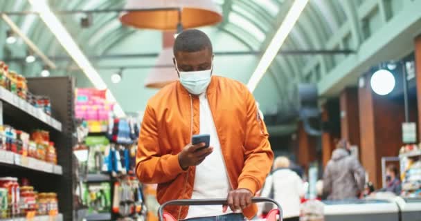 Homem afro-americano bonito em máscara protetora médica verificando lista de compras no supermercado. Jovem cliente masculino usando smartphone e comprando produtos na mercearia. Vírus, conceito pandémico. — Vídeo de Stock