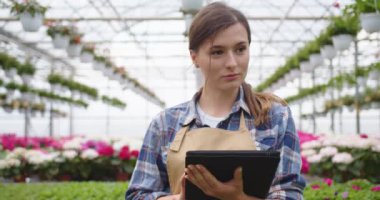 Close up of Caucasian young happy woman florist working in own floral shop walking in greenhouse and typing on tablet browsing online. Flower store owner. Garden center, small business concept