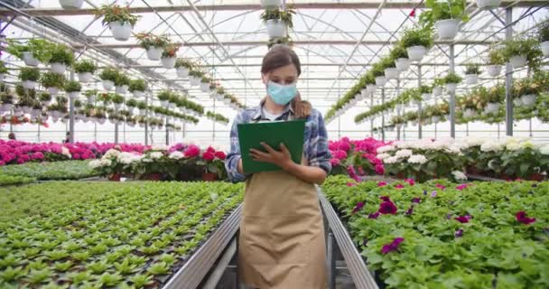 Mujer guapa joven caucásica gerente en máscara caminando en el centro del jardín y escribiendo usando portapapeles trabajando en su propia tienda de flores. Invernadero, tienda floral, cuarentena, concepto de negocio — Vídeos de Stock