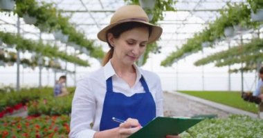 Close up portrait of cheerful young Caucasian woman gardener in apron and hat standing in greenhouse writing in clipboard, checking plants while walking. Garden center, floral shop. Floristry concept