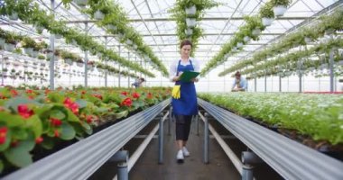 Happy beautiful young Caucasian woman in blue apron walking in own big greenhouse store writing in clipboard, checking plants. Gardener job, botanical. Small floral business concept