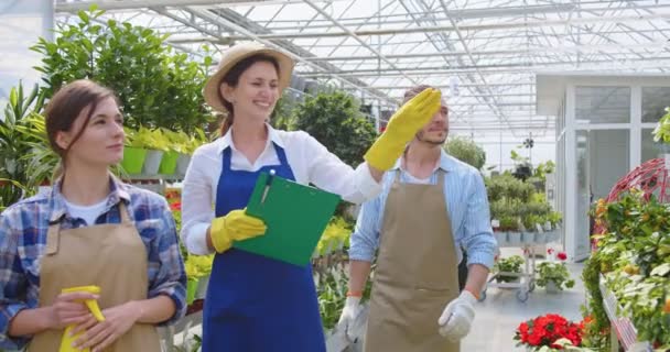 Portret van de blanke vrolijke vrouwelijke tuinman serre eigenaar spreken met jonge mannelijke en vrouwelijke werknemers in schorten tonen planten en bloemen binnen. Mensen die in het tuincentrum werken — Stockvideo