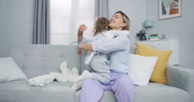 Güzel annenin sarışın kıvırcık kızını kucaklayıp modern oturma odasındaki kanepede oturmasının orta planı. Mutlu anne, tatlı küçük çocukla eğleniyor. Aile kavramı, ebeveynlik, birliktelik..