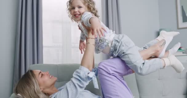 Plan medio de mamá sonriente jugando con un niño pequeño y adorable sosteniendo a un niño lindo, divirtiéndose, vuela en brazos de mamá haciendo ejercicio de gimnasia en el sofá. Concepto de familia, paternidad, unidad. — Vídeos de Stock