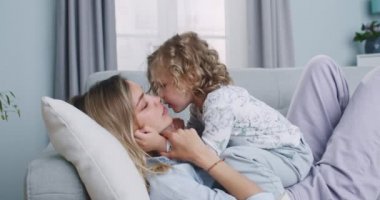 Middle plan of cute adorable funny little daughter caressing young mum, kissing on cheek, playing on sofa at home. Happy family having fun on couch. Concept of family, parenthood, togetherness.