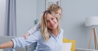 Close up of beautiful mum playing piggybacking with sweet kid daughter sitting on couch at home in beautiful living room, cheerful mother carrying small child girl on back bonding pretending flying.