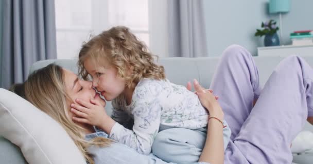 Plano medio de dulce niño divertido cosquillas mamá joven, besándose en la mejilla, jugando en el sofá en casa. Madre e hijo pequeño disfrutando de la unión en el sofá. Concepto de familia, paternidad, unidad. — Vídeo de stock