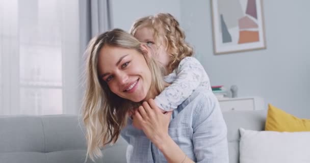 Close up of funny adorable cute little daughter kid hug caressing young mum. Kissing and playing on sofa. Happy family concept. Mother and small child girl enjoying and having fun on couch. — Stok video