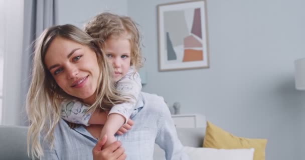 Close up of the beautiful caucasian mother and daughter on the sofa hugging and looking at camera. Happy mom having fun together with cute little child. Concept of family, parenthood, togetherness. — Stok video