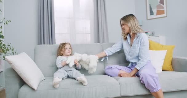 Середній план няні або няні, що тримає кролика, розважається з милою дитиною, що сидить на дивані, матір'ю і маленькою дівчинкою, яка грає ляльок разом. Концепція сім'ї, батьківства, взаємопорозуміння . — стокове відео