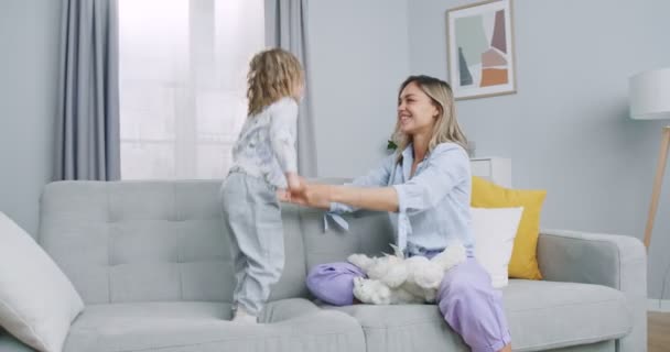Middle plan of cute active little girl jumping on comfortable furniture sofa having fun with mom at home, happy adult mother playing with small child daughter enjoy game on couch holding toys. — Stok video