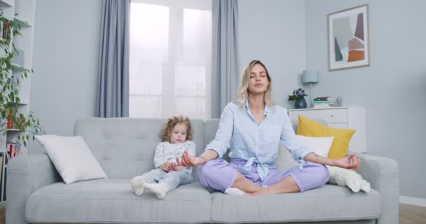 Piano di mezzo di triste figlioletta seduta accanto alla madre mentre la mamma medita in loto posa a casa. Felice sana madre di famiglia che fa esercizio di yoga rilassante seduta sul divano con figlia. — Video Stock