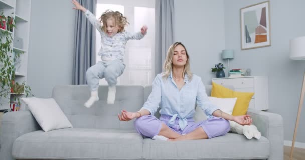 Consciente madre soltera meditando sentada en el sofá mientras la hija activa y enérgica salta jugando, calma joven mamá haciendo ejercicio de yoga en casa para aliviar el estrés relajándose con el niño travieso — Vídeos de Stock