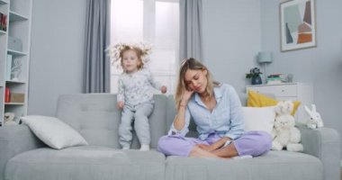Middle plan of tired mother meditating sitting on sofa while active energetic daughter jumping playing, sad young mom doing yoga exercise at home for stress relief relaxing with naughty little kid.