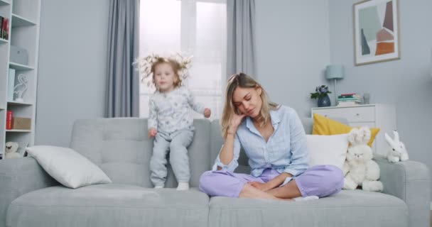 Plan medio de madre cansada meditando sentada en el sofá mientras activa hija enérgica saltando jugando, triste mamá joven haciendo ejercicio de yoga en casa para aliviar el estrés relajándose con el niño travieso. — Vídeos de Stock