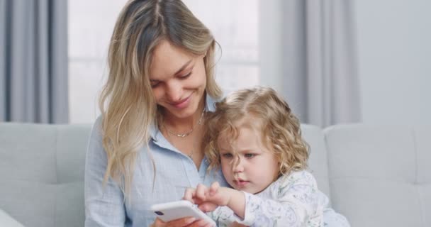 Középső terv aranyos vicces kisgyerek lány szórakozás anyával élvezze a modern szerkentyű okos telefon néz rajzfilmek kanapén otthon. A család fogalma, a szülőség, az összetartozás. — Stock videók