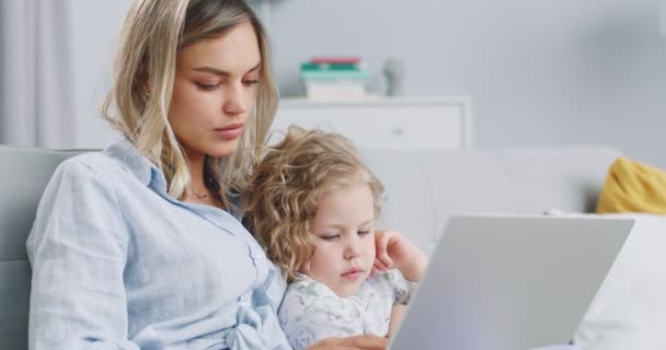 Mellanplan av attraktiv kvinna som arbetar på bärbar dator och kysser barn medan hennes vackra trevliga dotter sitter på soffan bredvid henne. Begreppet familj, föräldraskap, samhörighet. — Stockvideo