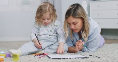 Middle plan of cute adorable kid girl holding color pencil learn drawing lying on warm floor with young mom daycare babysitter talking playing teaching helping little child daughter at home.