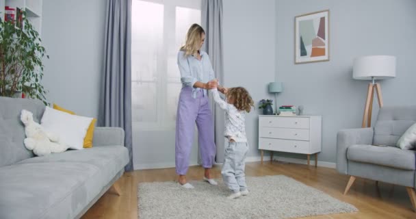 Plan moyen de baby-siter jeune maman active et mignonne petite fille sautant dansant dans le salon de la maison moderne, mère avec enfant fille s'amusant profiter de jouer à une activité drôle ensemble à la maison. — Video