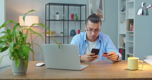 Genç Asyalı bir adam ofisteki bir masada dizüstü bilgisayarla çalışıyor. Kredi kartı bilgilerini akıllı telefonuna kaydediyor.