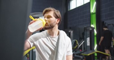 İçmek istiyorum. Omzunda havluyla bankta oturan spor sonrası rahatlarken aç gözlülükle içki içen fitness adamının belden yukarı portre görüntüsü. Spor ve eğlence.