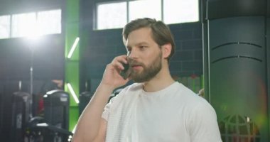 Neşeli adam spor salonunda dikiliyor ve elinde akıllı bir telefonla biriyle keyif sohbetinin tadını çıkarıyor. Egzersiz odasında çalışma arasında mola veriyor.