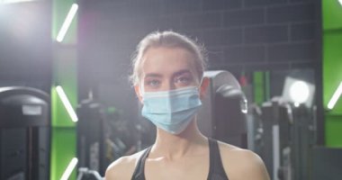 Karantina sırasında spor salonunda antrenman yaparken yüzünde koruyucu maskeyle kameraya bakan güçlü beyaz kadının portre görüntüsü. Covid 19 konsepti