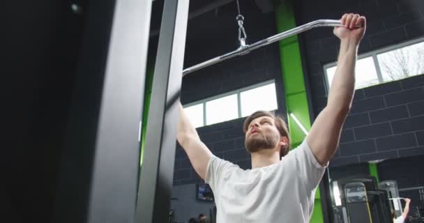 Trx fitnessapparaat in de sportschool. Gevoelig zicht op de actieve kalme sterke man trainingsarmen in activiteitencentrum met een plezierige glimlach met de sportschool met mensen op de achtergrond — Stockvideo