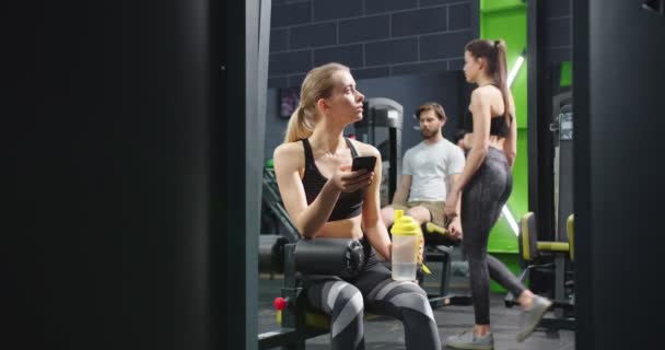 Full längd syn på fitness kvinnan i sportkläder sitter på bänken med flaska vatten och använda smartphone medan du tittar på video om sport. Idrott och fritid — Stockvideo