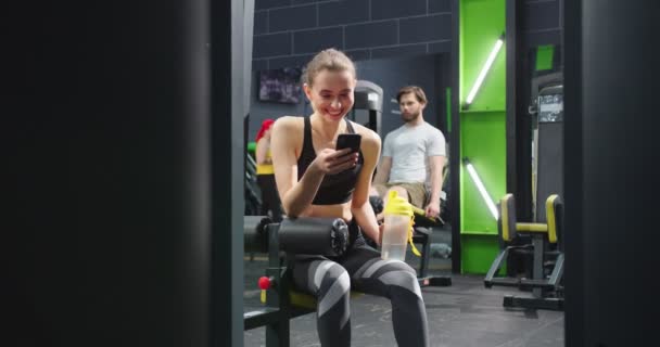 Vacker ung kaukasisk kvinna koppla av efter träning och dricksvatten från plastflaskan medan du tittar på fitness videor och ler mot gymmet. Idrott och fritid — Stockvideo