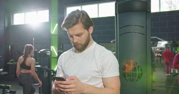 Forte atleta barbuto uomo è in piedi in palestra e riposo dopo un duro allenamento. Guy sta guardando lo schermo dello smartphone mentre tiene in mano il gadget. Video stock — Video Stock