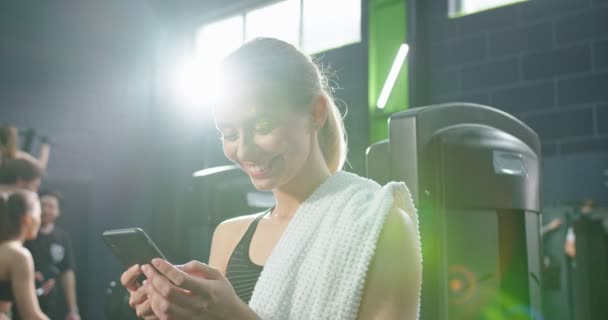 Ισχυρή αθλήτρια ελκυστική γυναίκα στέκεται στο γυμναστήριο και ξεκουράζεται μετά από σκληρή προπόνηση. Κορίτσι κοιτάζει οθόνη smartphone, ενώ κρατώντας gadget στο χέρι της. Απόθεμα βίντεο — Αρχείο Βίντεο