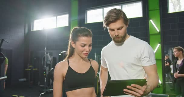 Tour de taille portrait de la fille sportive calme regardant quelque chose à l'écran de la tablette avec son entraîneur ou son entraîneur tout en constituant un programme d'entraînement. Concept de sport et de loisirs — Video