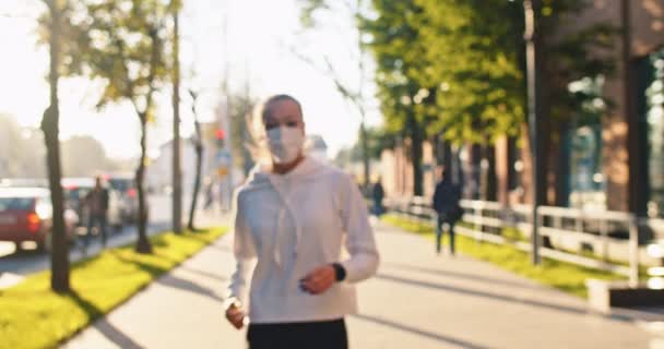 Close up of Caucasian young beautiful woman in mask running listening to music in wireless earphones in city on sunny street stop and removing mask from face. Fitness, outdoor training, exercising — Stock Video