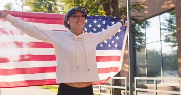 Porträt kaukasischer junger glücklich lächelnder, fitter Frauen, die auf der Straße mit einer US-Fahne laufen. Fröhliche Frau mit amerikanischer Flagge im Freien an einem sonnigen Sommertag beim Sport. Outdoor-Training, Outdoor-Konzept — Stockvideo