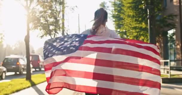 Tył młodej białej kobiety sportowej biegającej po słonecznej ulicy z flagą USA. Kobieta z amerykańską flagą biegająca w letni dzień. Trening sportowy, ćwiczenia na świeżym powietrzu, koncepcja fitness — Wideo stockowe