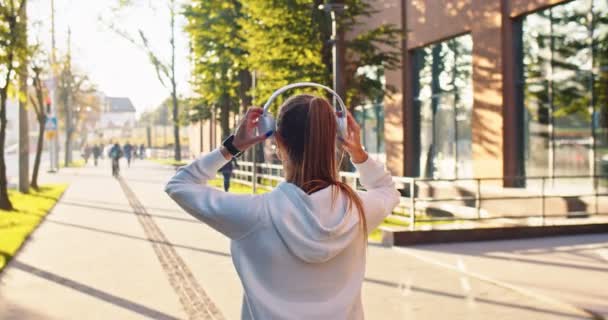 Visão traseira da jovem mulher andando ao ar livre na rua colocando fones de ouvido e ouvindo música de bom humor. Mulher caucasiana ter andar na cidade no dia ensolarado apreciando música, conceito traseiro — Vídeo de Stock