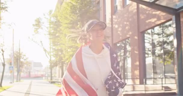 Porträt einer fröhlichen kaukasischen jungen lächelnden schönen Frau, die mit einer US-Fahne auf der Straße läuft und über Kopfhörer Musik hört. Fröhliche Frau mit amerikanischer Flagge joggt an sonnigen Tagen Sport-Workout — Stockvideo