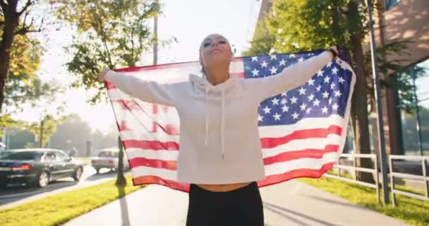Porträt einer fröhlichen positiven kaukasischen jungen hübschen Frau, die auf der sonnigen Straße läuft und joggt und die USA-Flagge hält. Glückliche Sportlerin mit amerikanischer Flagge im Freien an einem Sommertag. Sportkonzept — Stockvideo