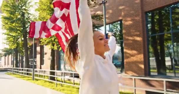 Großaufnahme einer jungen, fröhlichen Sportlerin aus dem Kaukasus, die auf der sonnigen Straße in der Stadt mit der US-Flagge läuft. Fröhliche Läuferin mit amerikanischer Flagge beim Joggen an einem Sommertag. Sport, Sport, Bewegung — Stockvideo