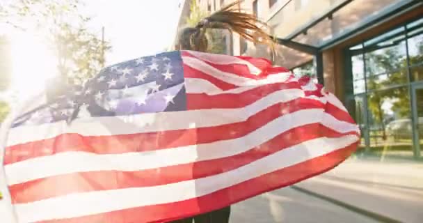 Visão traseira da jovem esportista caucasiana correndo na rua ensolarada na cidade segurando bandeira dos EUA. Desportista com bandeira americana correndo no dia de verão. Estilo de vida desportivo, treino, conceito de fitness — Vídeo de Stock