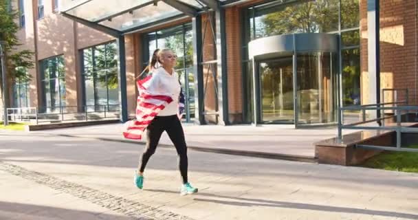 Fröhliche kaukasische junge hübsche Läuferin joggt draußen in der Stadt und lächelt mit amerikanischer Flagge darüber. Glücklich schöne, fitte Hündin mit USA-Fahne, die nach draußen läuft. Sonniger Tag, patriotisches Konzept — Stockvideo