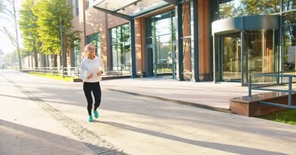Caucasian young beautiful happy sportswoman running in sunny town in good mood, morning exercises. Joyful pretty female jogging outdoors listening to music in earphones. Sport lifestyle concept — Stock Video