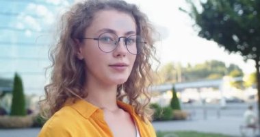 Kameraya bakıp gülümseyen gözlüklü, mutlu, genç beyaz kadını yakından çek. Dışarıda poz veren kıvırcık saçlı güzel bir kadın. Açık hava kavramı