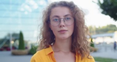 Kamera, gözlüklü, güzel, beyaz, mutlu bir bayana yaklaşıyor. Şehirde durmuş, yüzünde gülümsemeyle kameraya bakıyor. Kasabada gülümseyen kıvırcık saçlı güzel, neşeli bir kadın. Portreyi kapat