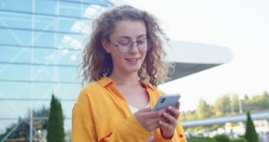 Kasabada sokakta yürüyen ve akıllı telefondan mesajlaşan gülümseyen beyaz, genç ve güzel bir kadının portresini yakından çek. Sosyal ağ uygulaması kullanarak cep telefonunda yazan neşeli, güzel bir kadın.