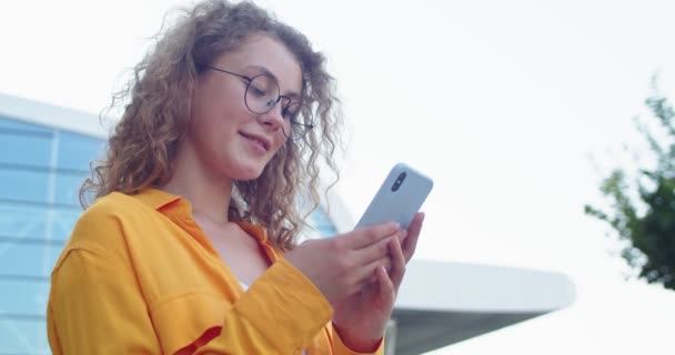 Κοντινό πλάνο του Καυκάσου νεαρή όμορφη χαρούμενη γυναίκα με γυαλιά στέκεται στο δρόμο και γραπτών μηνυμάτων σε smartphone με χαμόγελο στο πρόσωπο. Ευτυχισμένο όμορφο θηλυκό περιήγηση σε απευθείας σύνδεση χρησιμοποιώντας την εφαρμογή κοινωνικής δικτύωσης στην πόλη — Αρχείο Βίντεο
