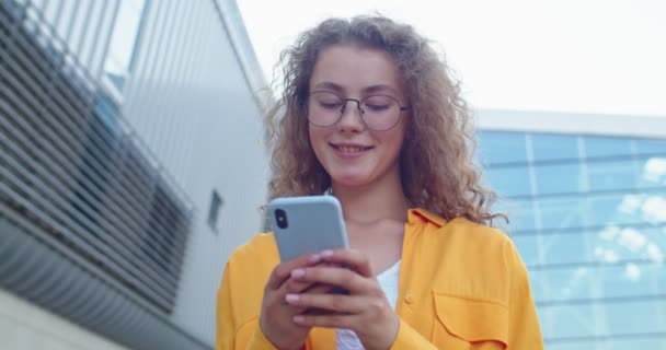 Κοντινό πλάνο πορτρέτο του Καυκάσου νεαρή ευτυχισμένη σγουρά γυναίκα σε γυαλιά στέκεται στο δρόμο στην πόλη και γραπτών μηνυμάτων σε smartphone Χαρούμενη όμορφη γυναίκα δακτυλογράφηση σε απευθείας σύνδεση σε εφαρμογή κοινωνικής δικτύωσης, αστικό στυλ — Αρχείο Βίντεο