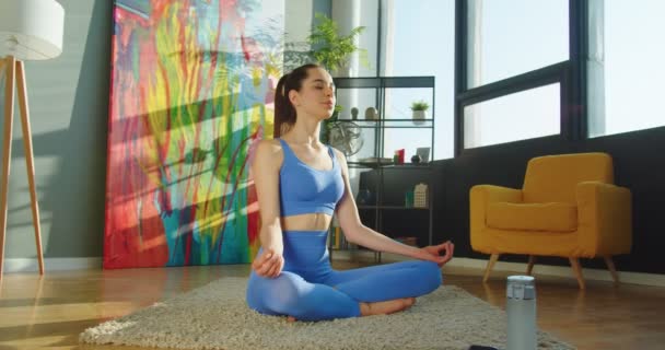 Hermosa joven deportista feliz en ropa deportiva sentado en el suelo de la alfombra y meditando en la habitación soleada. Mujer atlética caucásica practicando yoga en casa, cuerpo fuerte, deporte y meditación, relajación — Vídeos de Stock