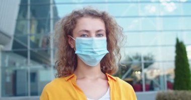 Sağlık maskeli beyaz bir kadının kameraya bakarak dışarıda neşeli bir şekilde durmasını yakın çekim yapın. Dışarıda poz veren kıvırcık saçlı güzel bir kadın. Covid salgını. Açık hava kavramı
